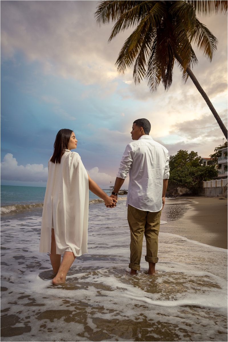 Couple holding hands on a tropical beach at sunset, standing in gentle waves near a palm tree.