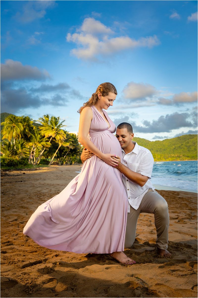 A couple shares an intimate moment on a tropical beach at sunset with palm trees and mountains in the background.