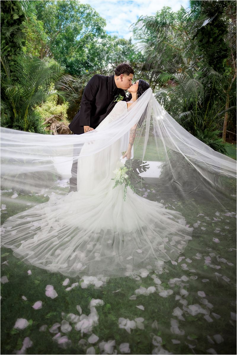 Long wedding veil flowing dramatically across a lush garden setting with scattered flower petals.