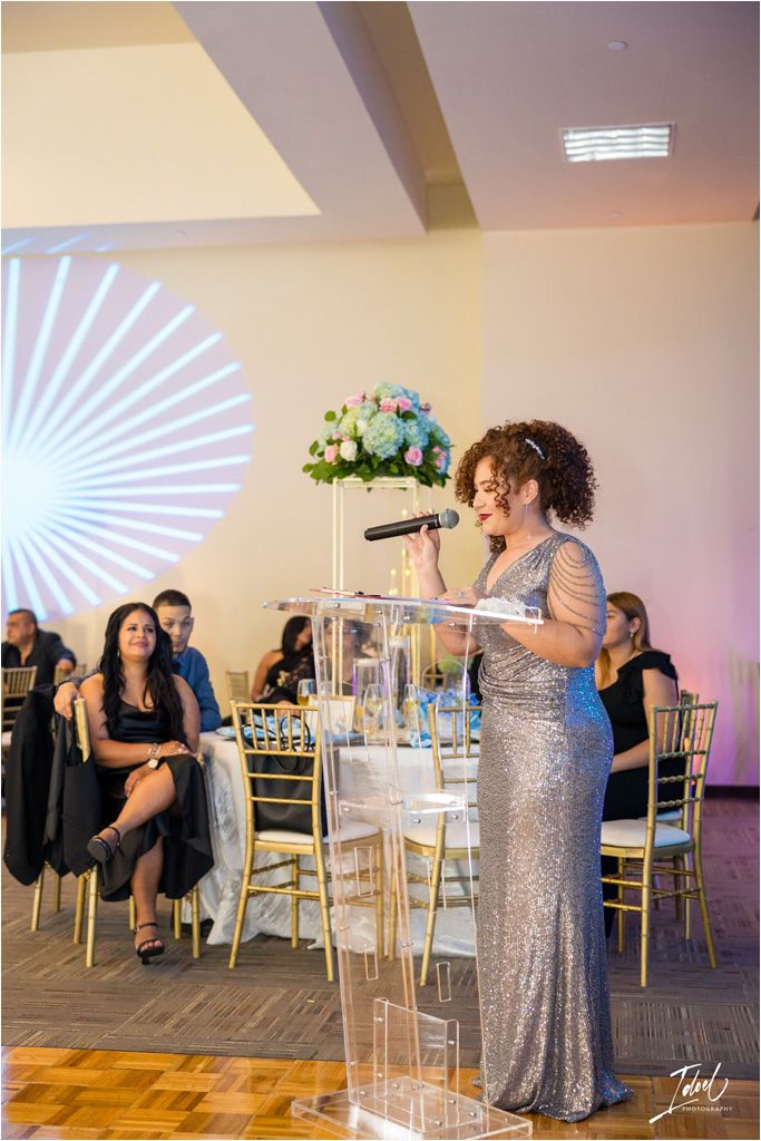A speaker in a sparkling gown delivers a speech at a wedding reception podium.