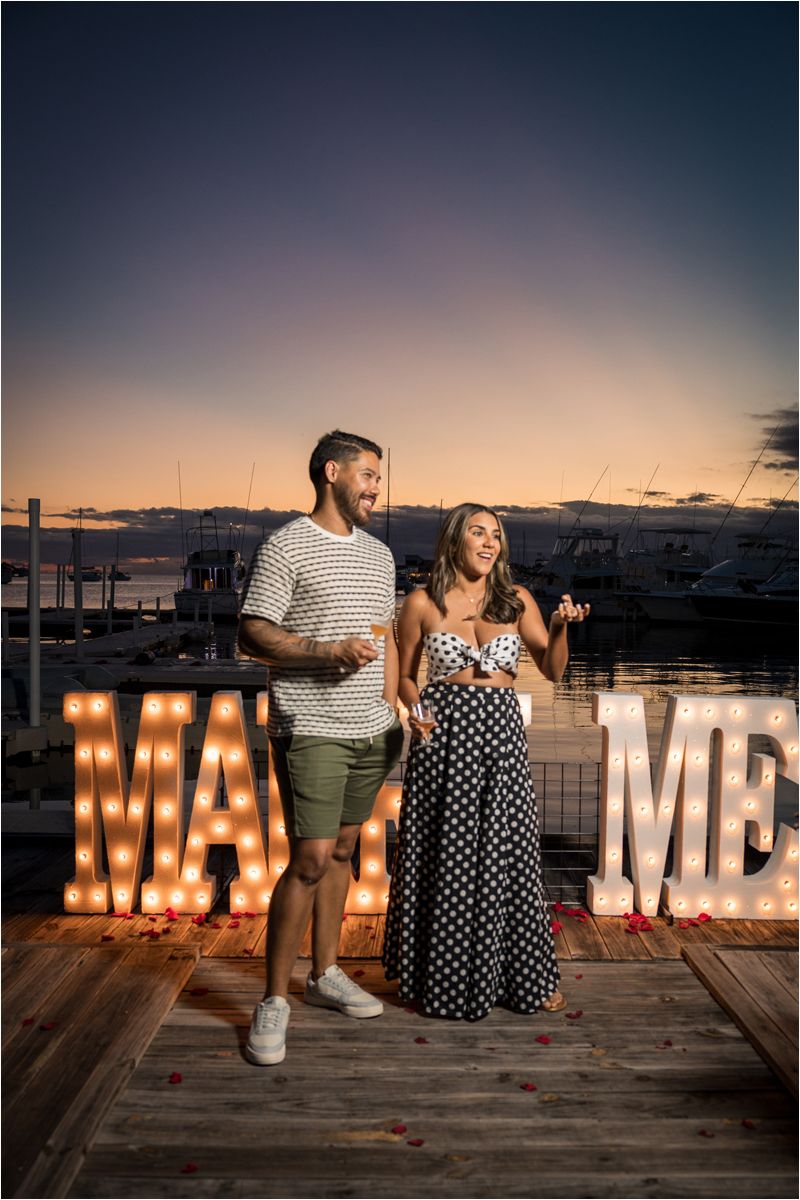 Couple stands on wooden deck at sunset with illuminated MARRY ME marquee letters creating romantic proposal ambiance.