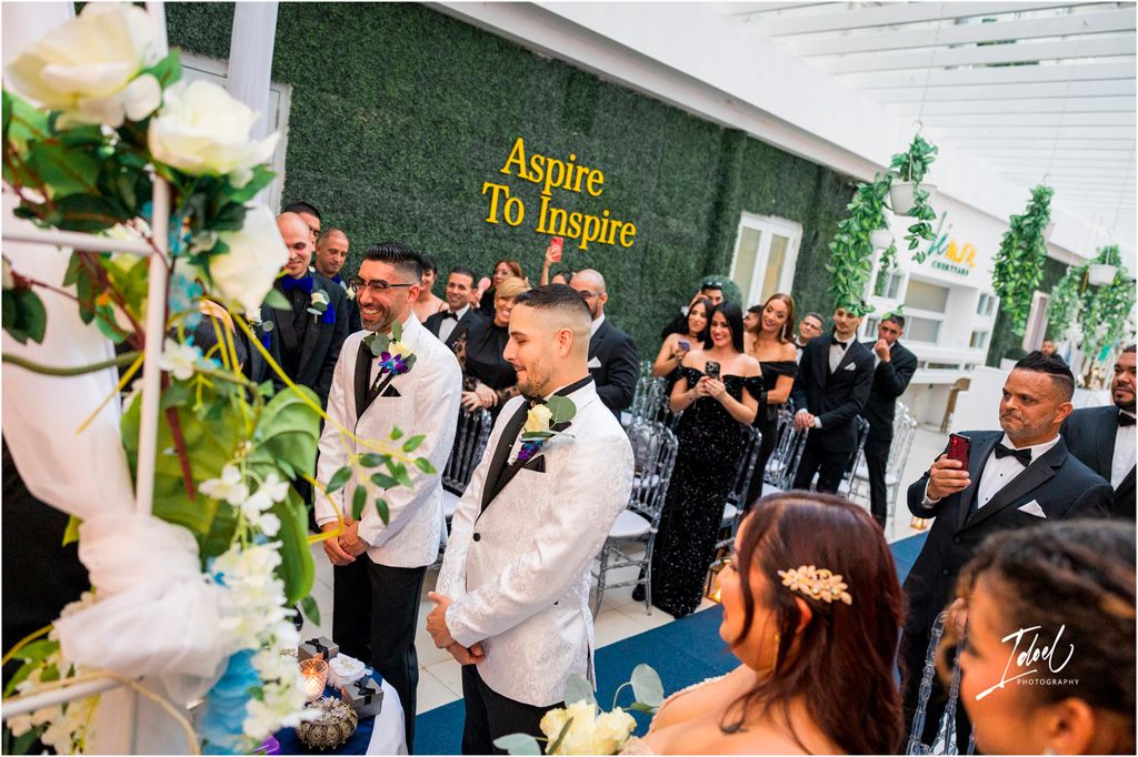 Wedding celebration features guests gathered near floral decorations and a green wall with gold lettering.
