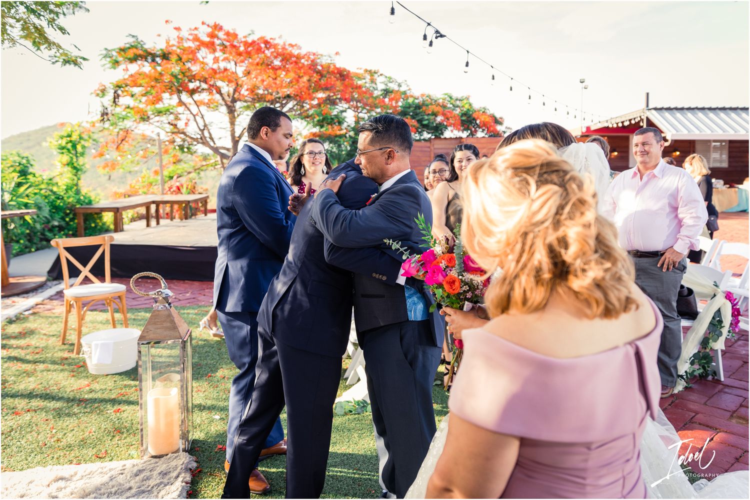Wedding guests mingling on a brick patio under string lights with a vibrant red tree in background.