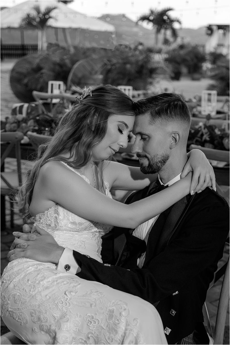 A romantic black and white portrait of a couple embracing outdoors, with the bride wearing a lace dress and groom in formal suit.