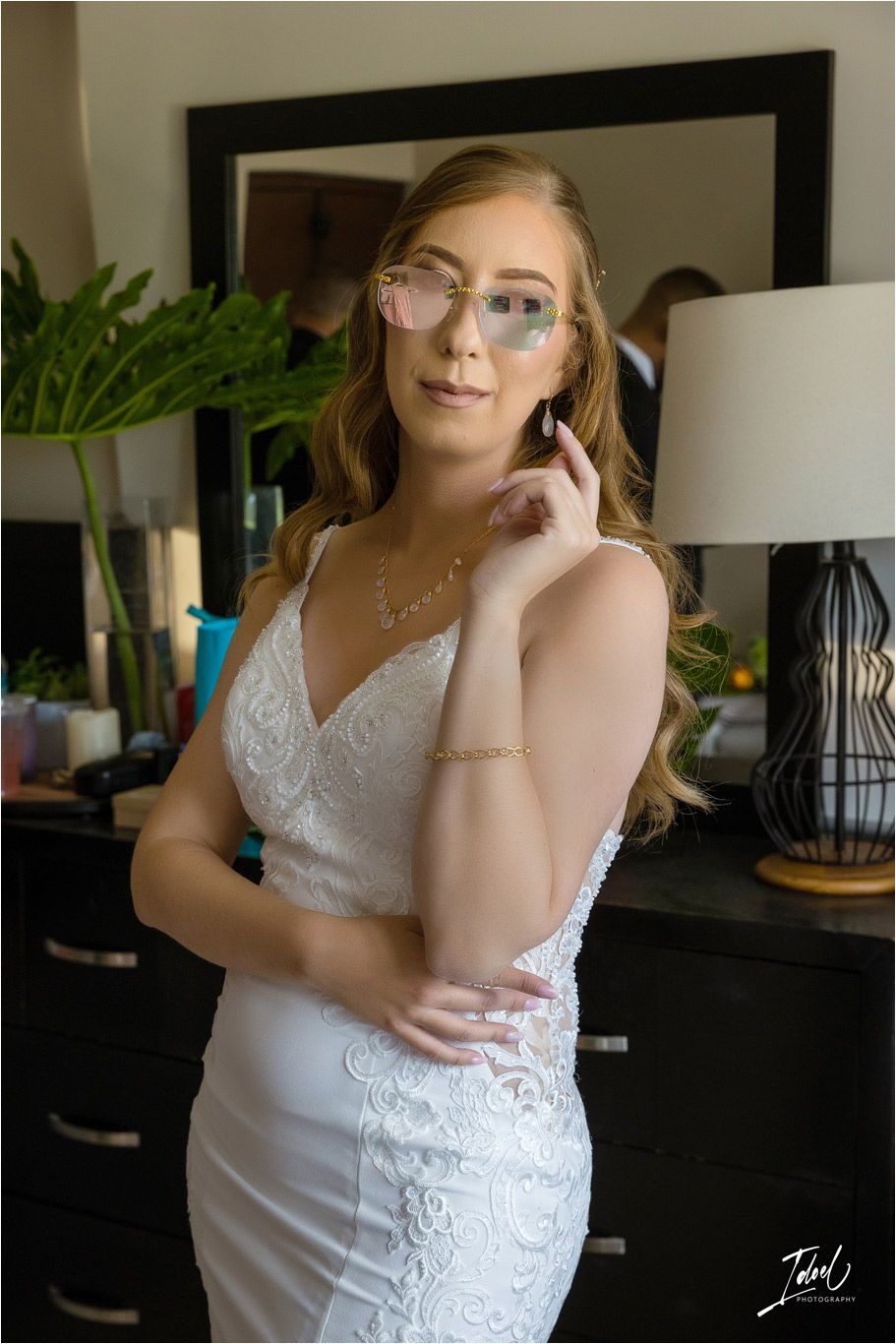 Woman in lace wedding dress posing elegantly next to a dresser with greenery decor.