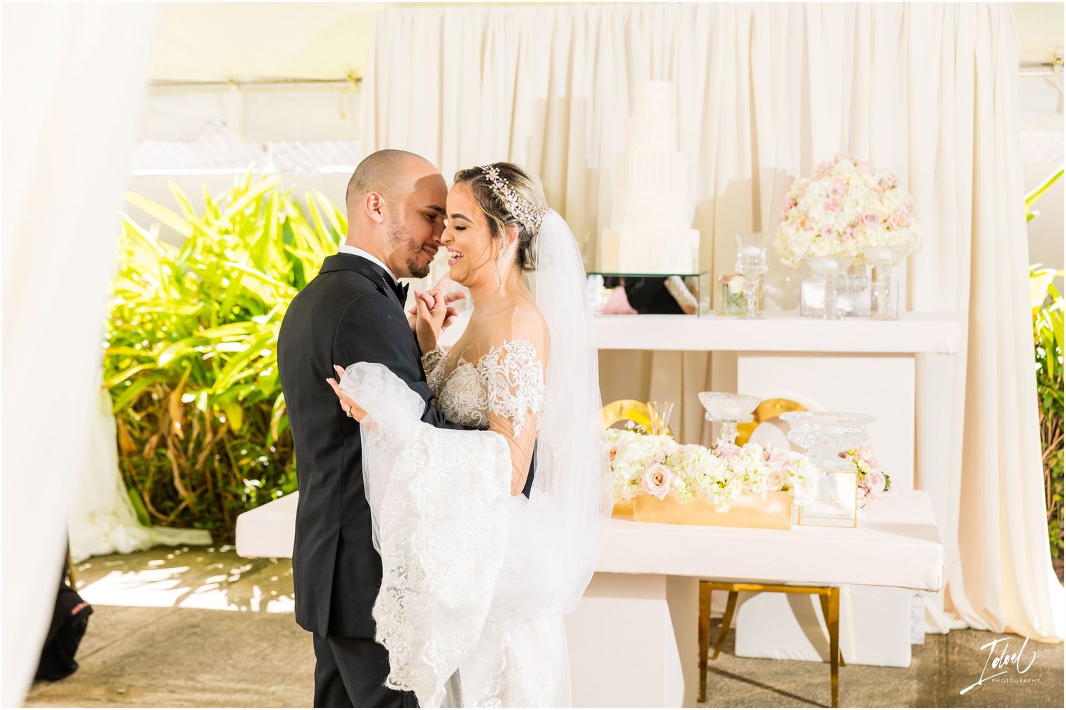 Wedding couple shares romantic moment under white tent with lush tropical garden backdrop.