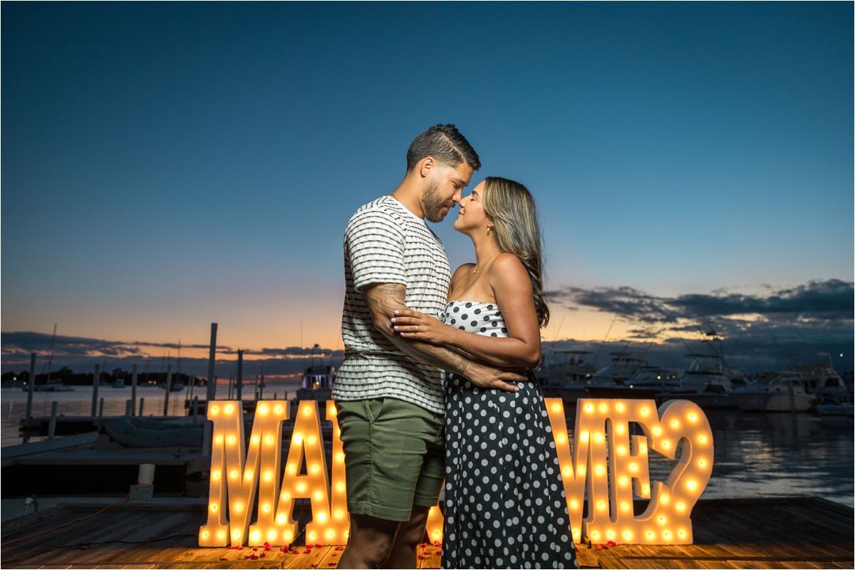 A couple embraces at sunset near illuminated marquee letters on a rooftop.