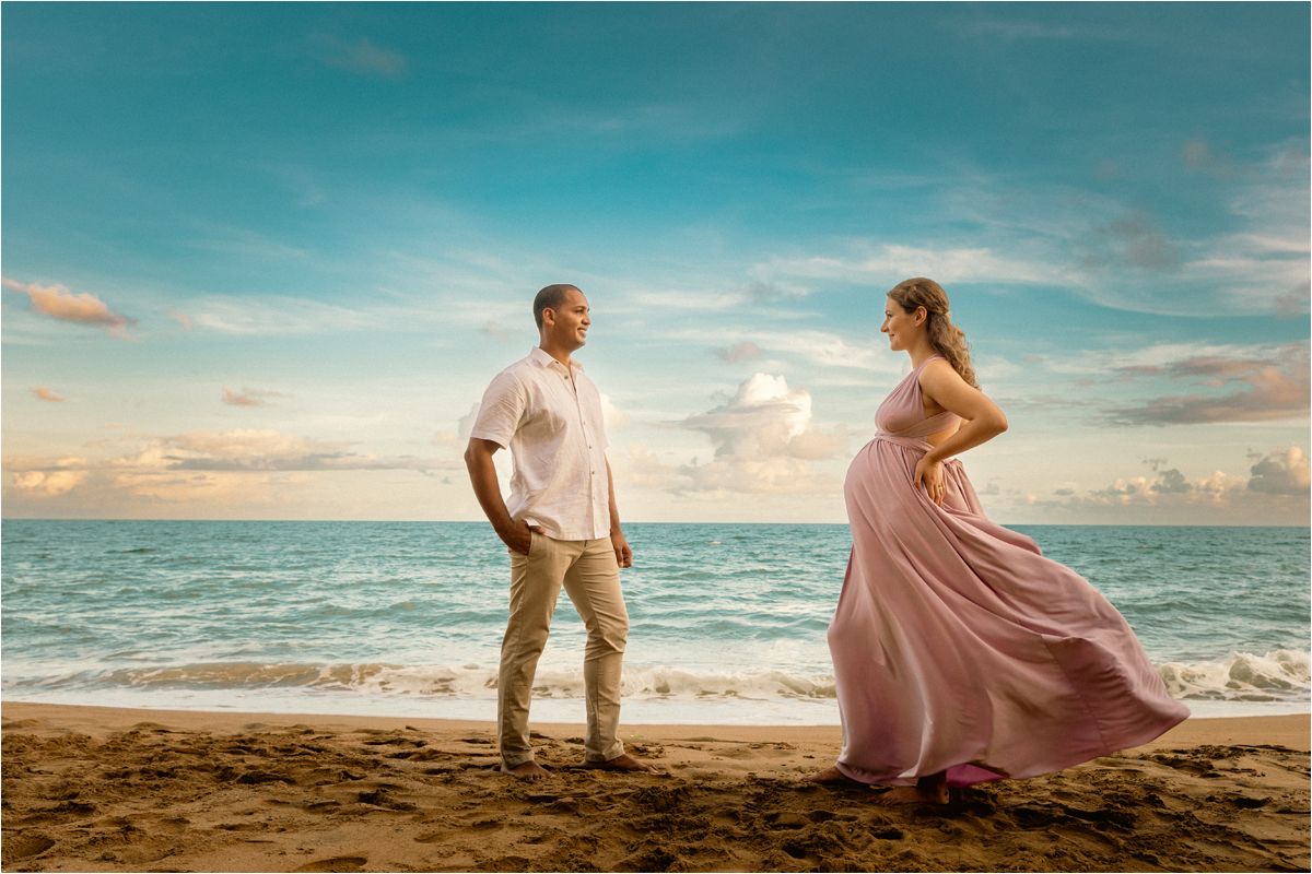 A couple in romantic attire stands on a sandy beach at sunset with waves crashing in the background.
