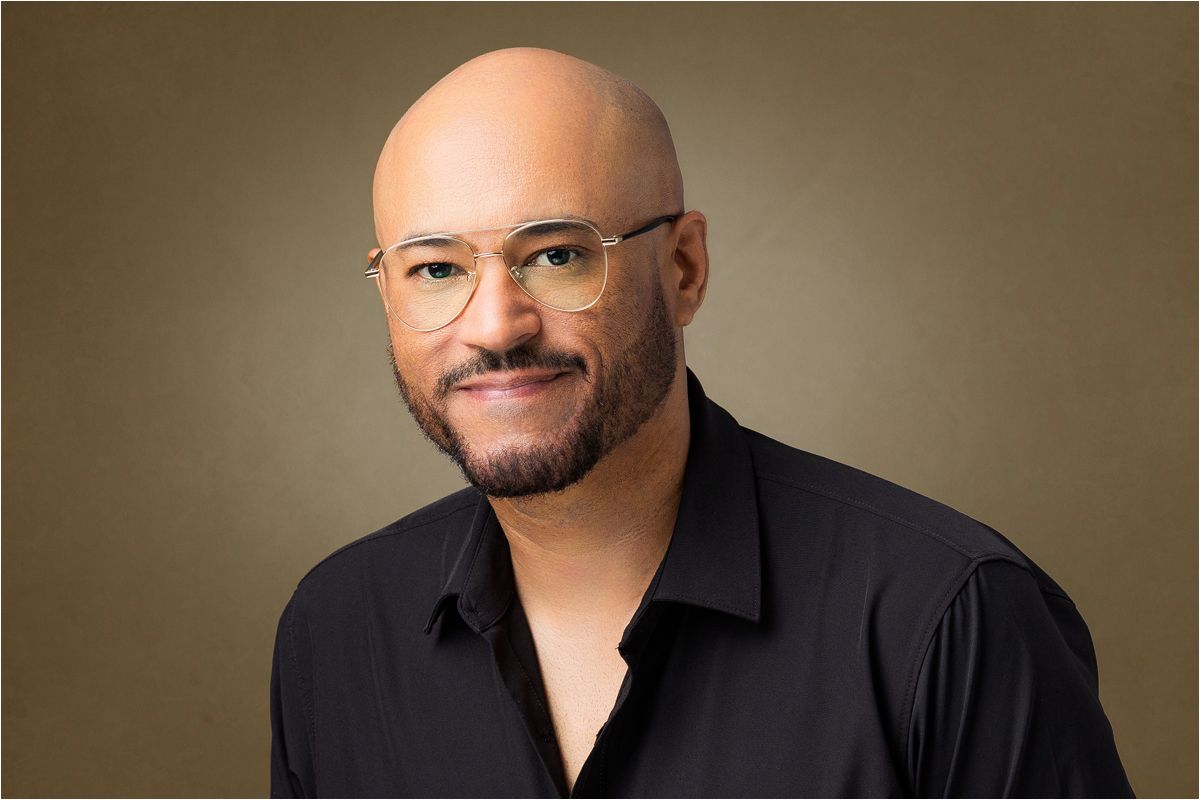 Professional headshot of a person wearing glasses and a black button-down shirt against a brown background.