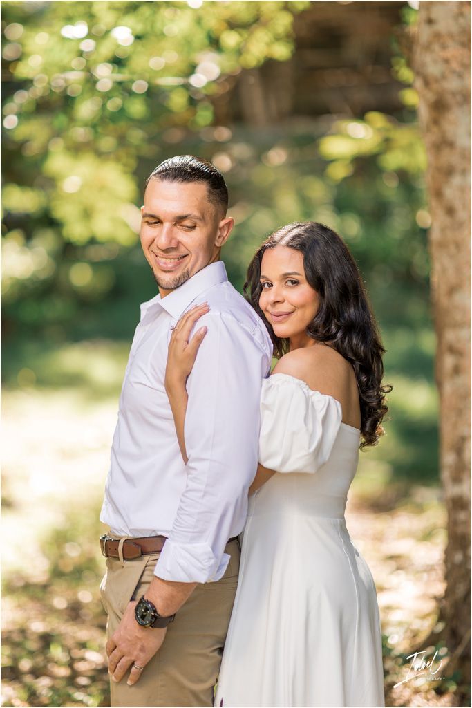 A couple poses together outdoors in romantic engagement photos with soft natural lighting filtering through trees.