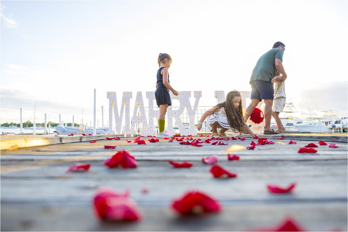 People collecting scattered red rose petals on a dock by the water during a romantic outdoor scene at sunset.