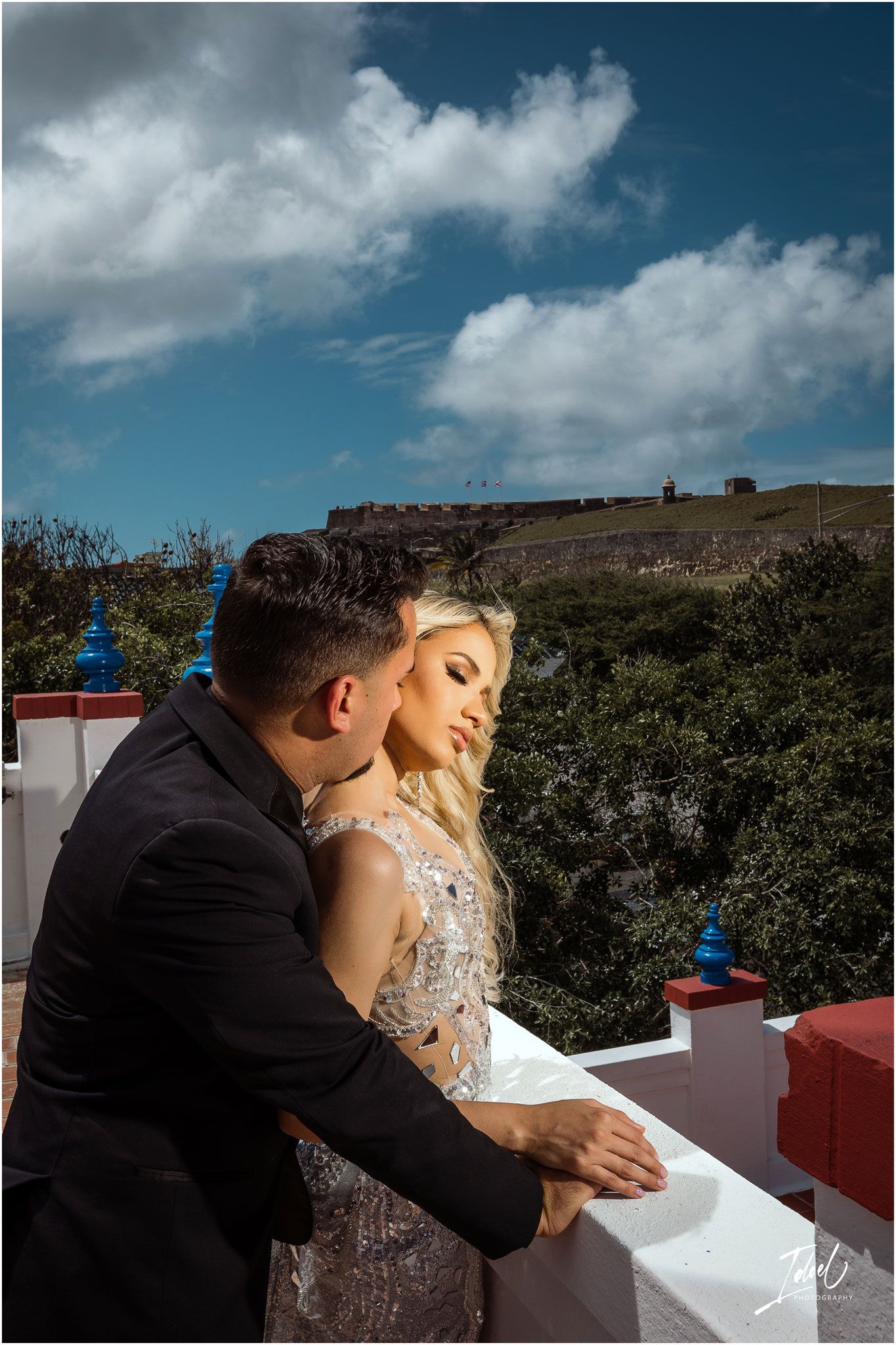 Romantic couple shares intimate moment on balcony with dramatic cloudy sky and fortress in background.