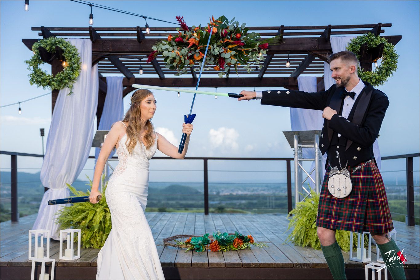 Couple duels with prop guns during playful wedding photoshoot on scenic outdoor deck.