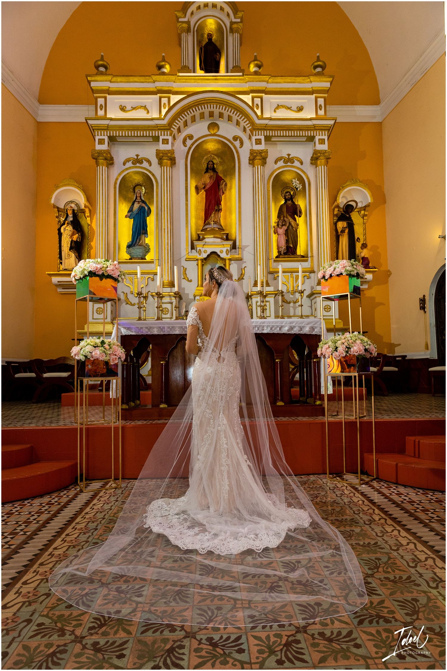Back view of bride in cathedral length veil standing before decorated church altar with saints statues.