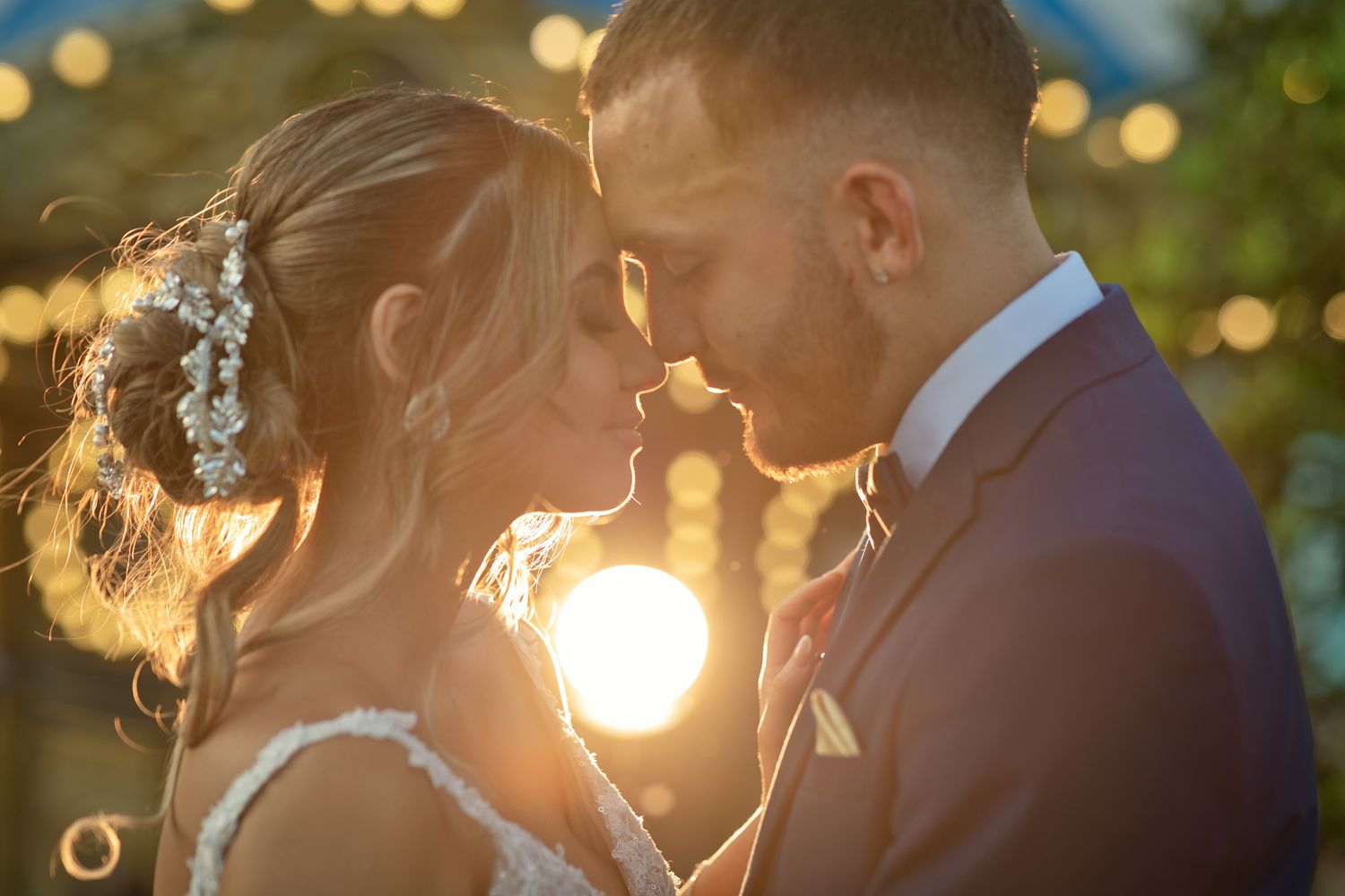 A bride and groom share a romantic sunset moment, foreheads touching, in golden backlit wedding photography.