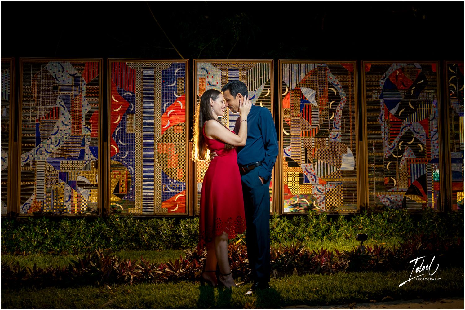 Couple embracing romantically at night in front of colorful mosaic mural, woman in red dress, man in blue suit.