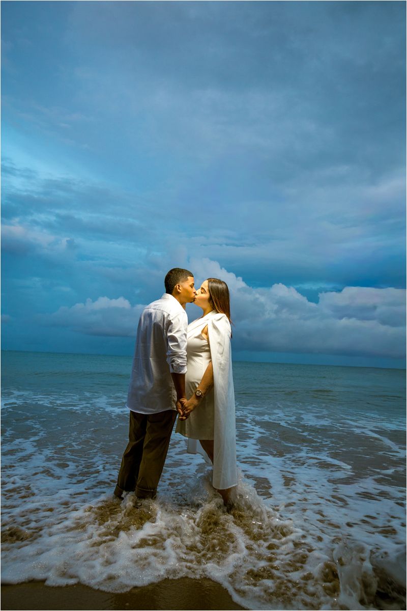 Couple kissing at the ocean's edge during twilight, waves washing over their feet, dramatic blue cloudy sky behind them.
