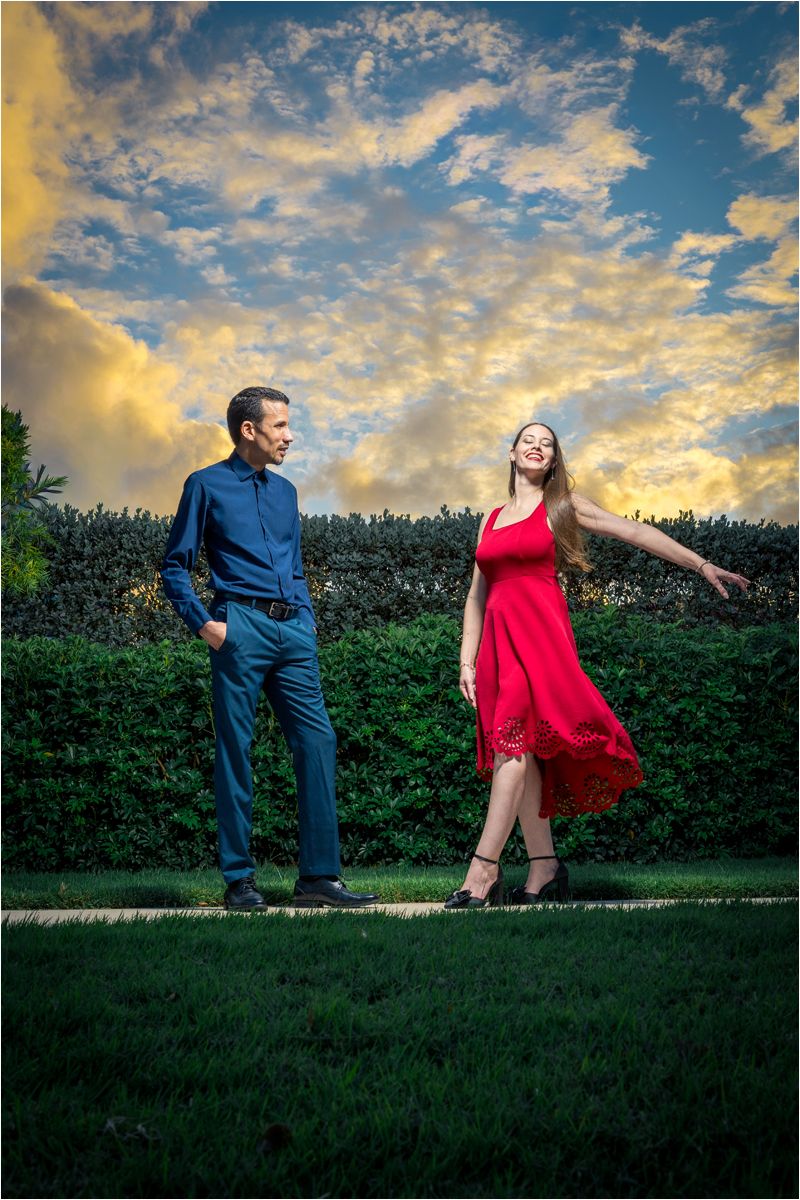 Couple posing outdoors, woman in red dress twirling, man in blue shirt, green hedge backdrop, dramatic cloudy sunset sky.