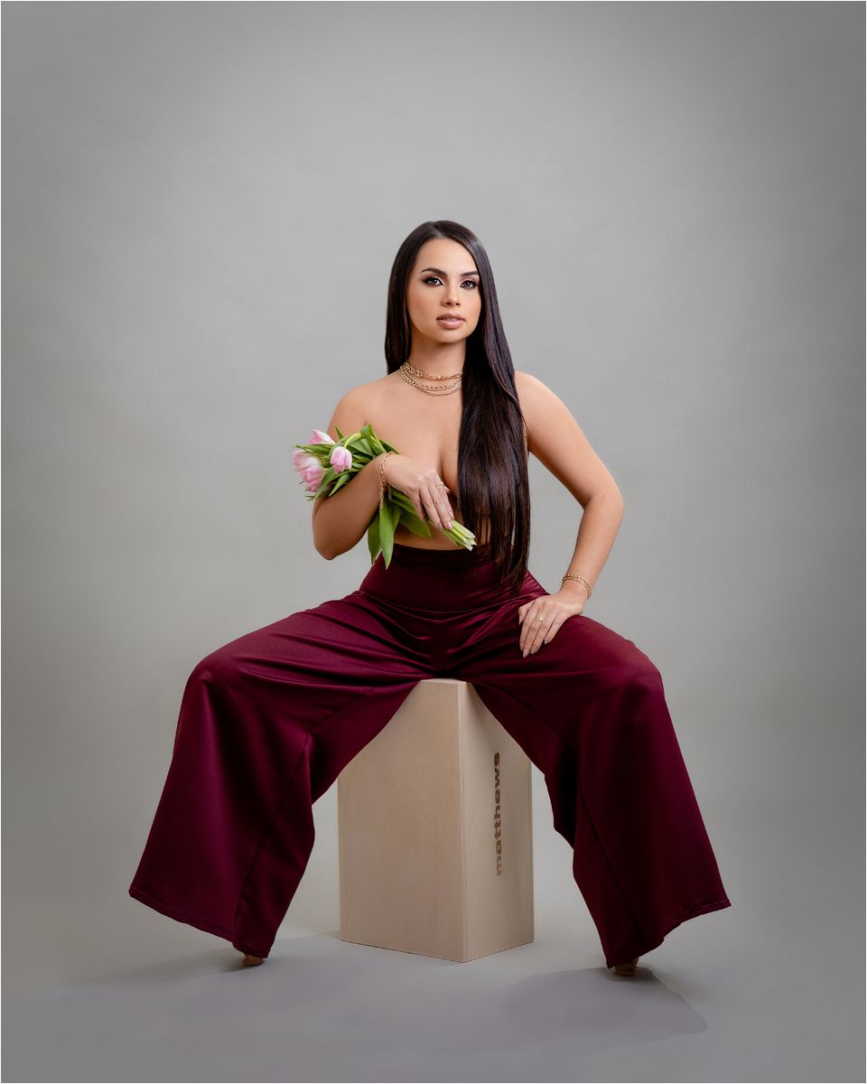 Woman in burgundy wide-leg jumpsuit poses confidently on minimalist cube against grey backdrop in fashion photoshoot.