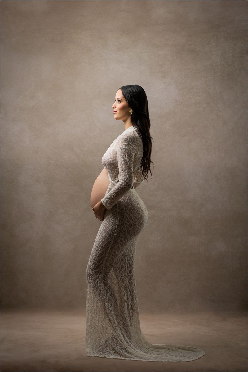 A side profile of an expectant mother in a long lace dress poses elegantly against a neutral studio backdrop.