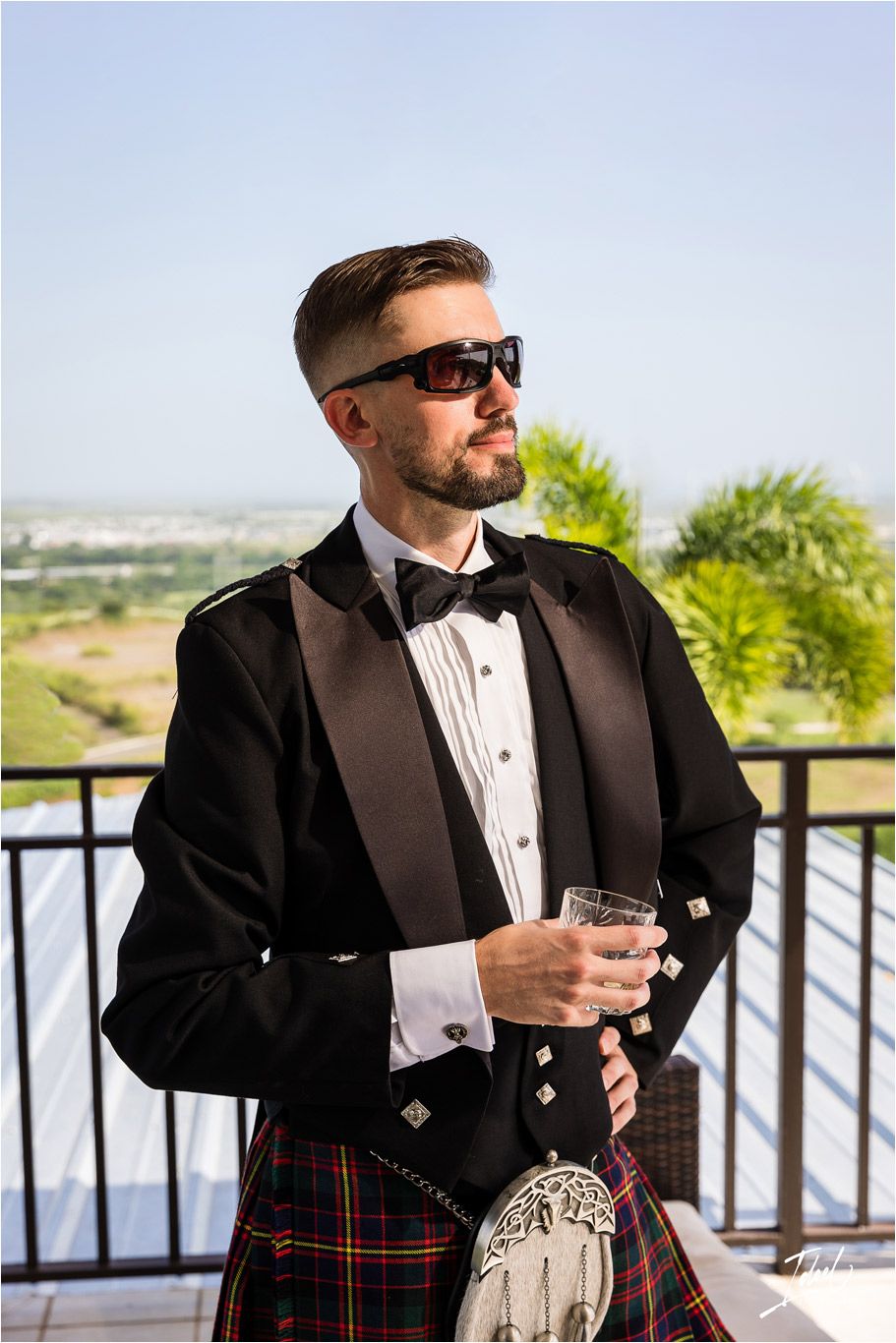 Groomsman wearing sunglasses and traditional Scottish formal wear poses with drink outdoors.