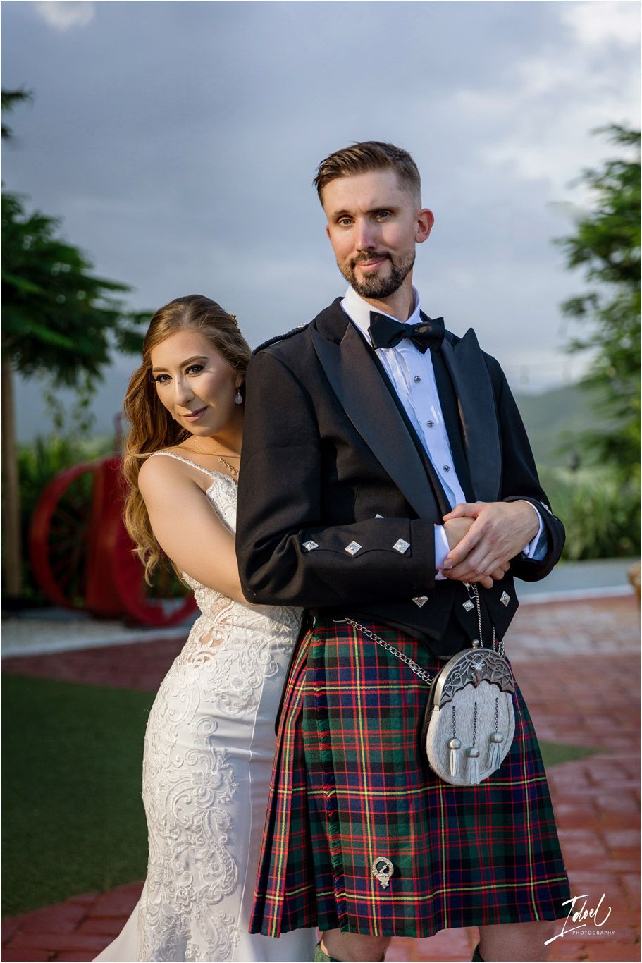 A couple poses in traditional Scottish wedding attire with a kilt and lace gown against a garden backdrop.