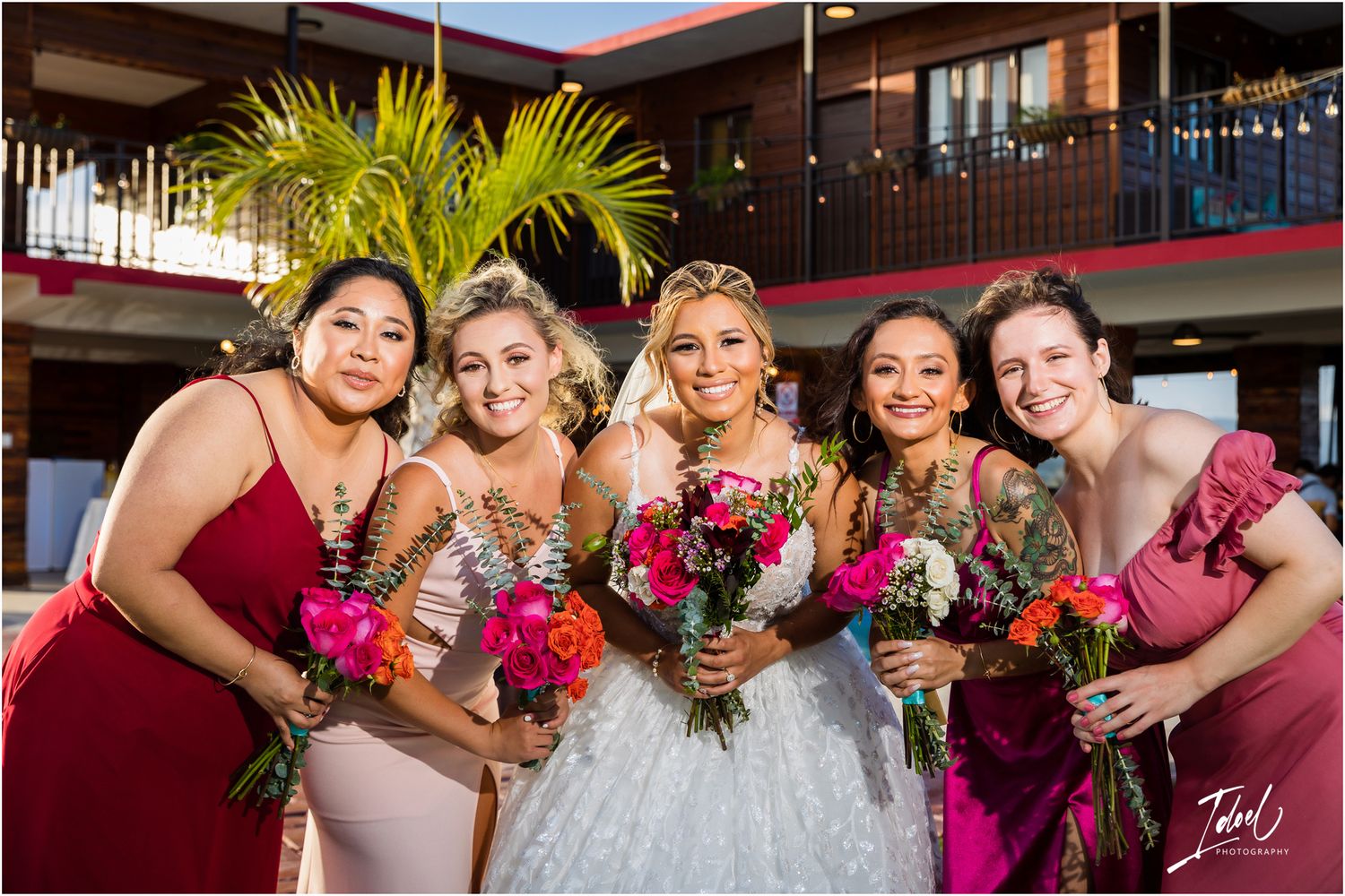 Bridal party in mix of burgundy and blush dresses hold vibrant pink bouquets outside tropical venue.