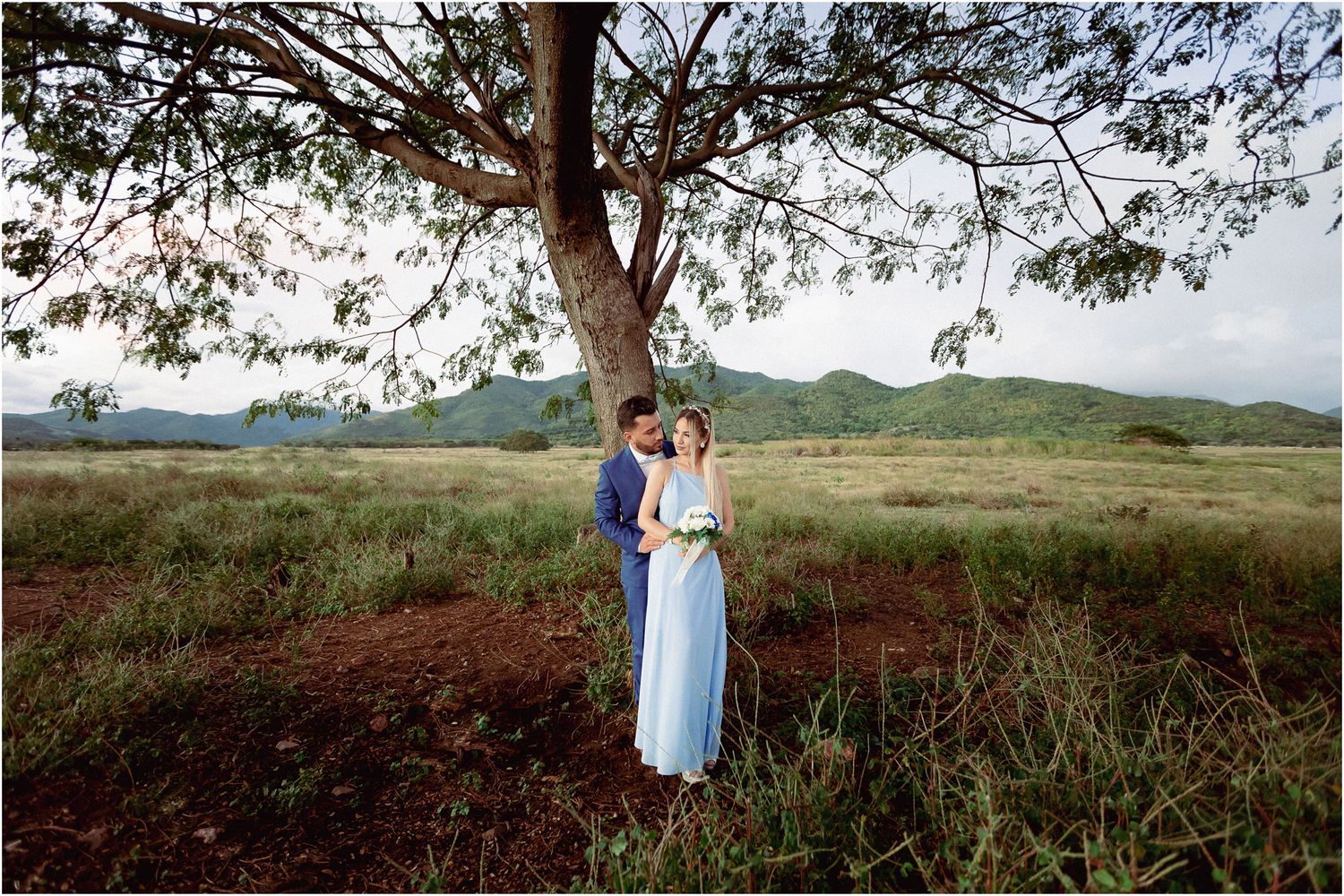 A couple shares a romantic moment under a large tree in a scenic rural landscape at sunset.