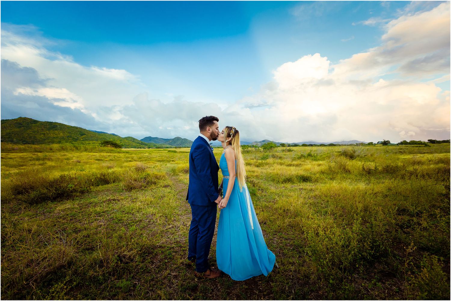 A couple in formal attire embraces in a sun-lit meadow with mountains visible in the background.
