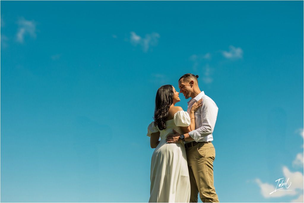 A romantic couple embraces against a vibrant blue sky with puffy white clouds on a sunny day.