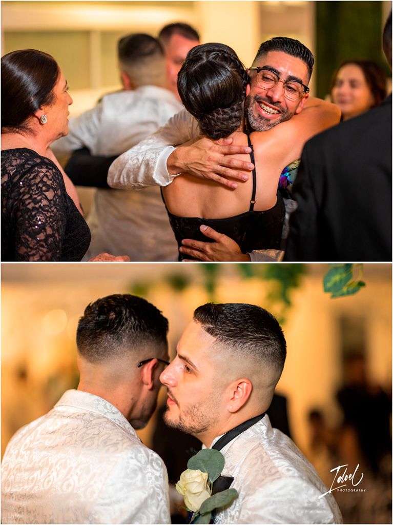 Two people in white dress shirts embrace warmly at a celebration while guests gather around them at a wedding reception.