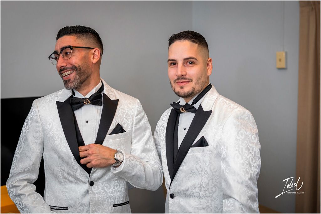 Two people wearing matching white floral tuxedos with black bow ties and lapels smile together at formal event.