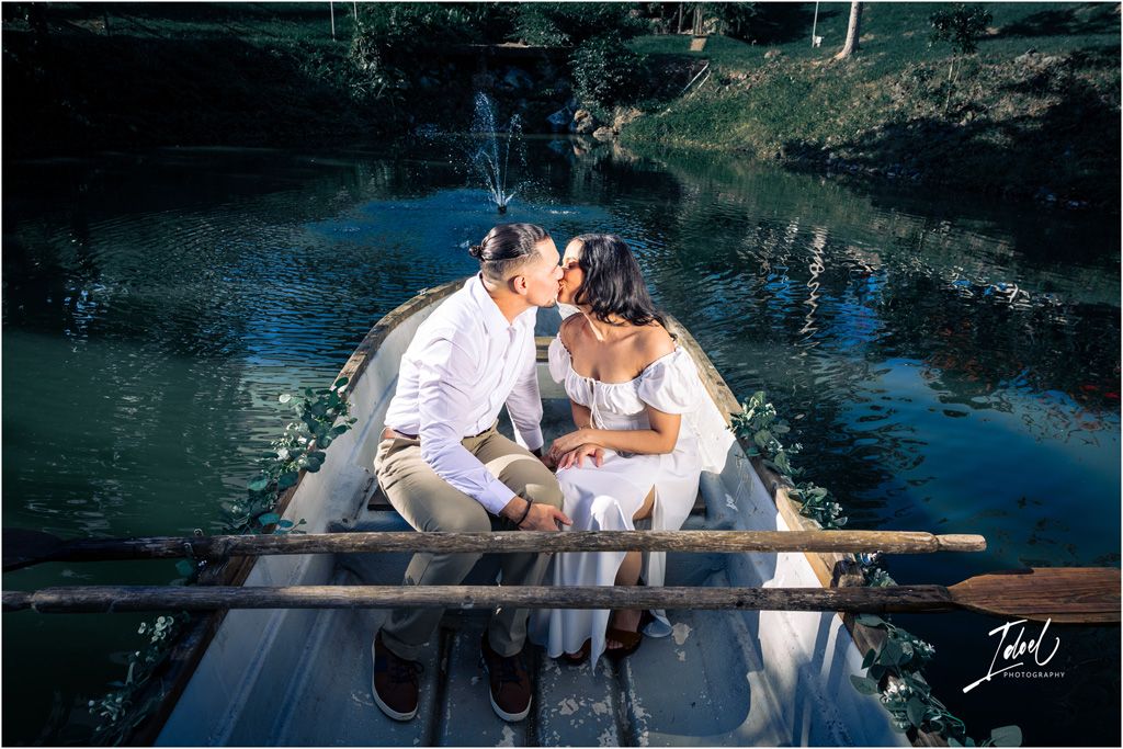 A romantic couple shares a kiss while sitting in a boat on a serene blue lake surrounded by greenery.