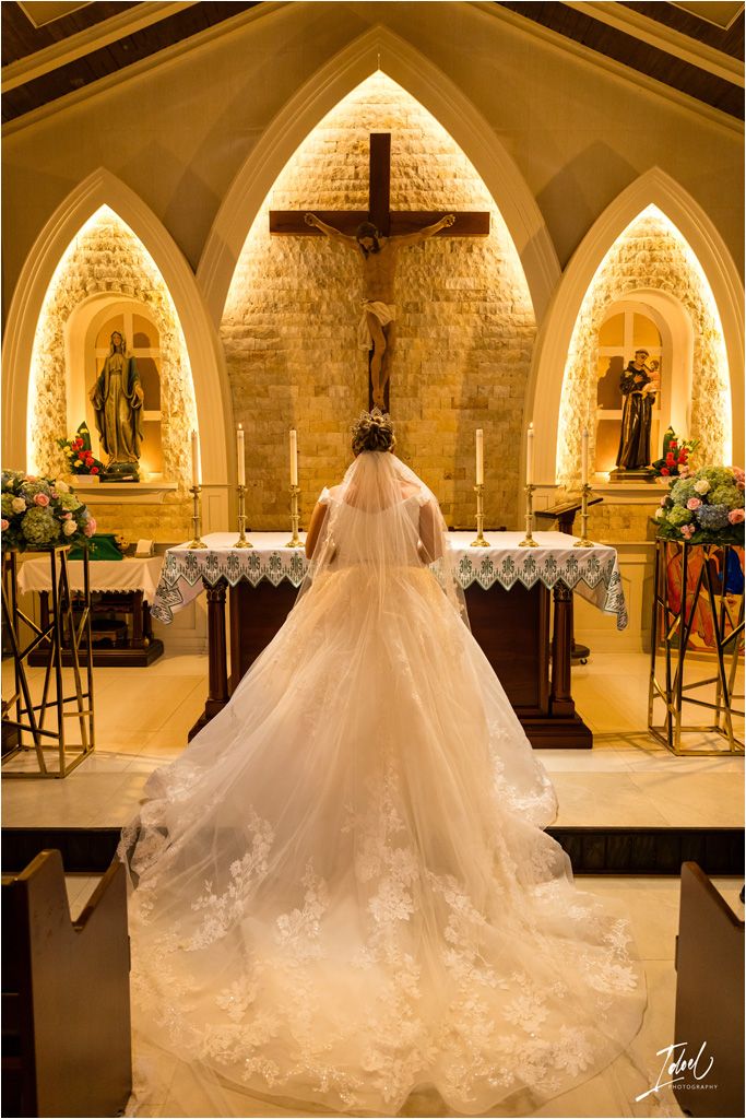 A bride in a flowing lace wedding gown stands before an illuminated altar with gothic arches and religious statues.
