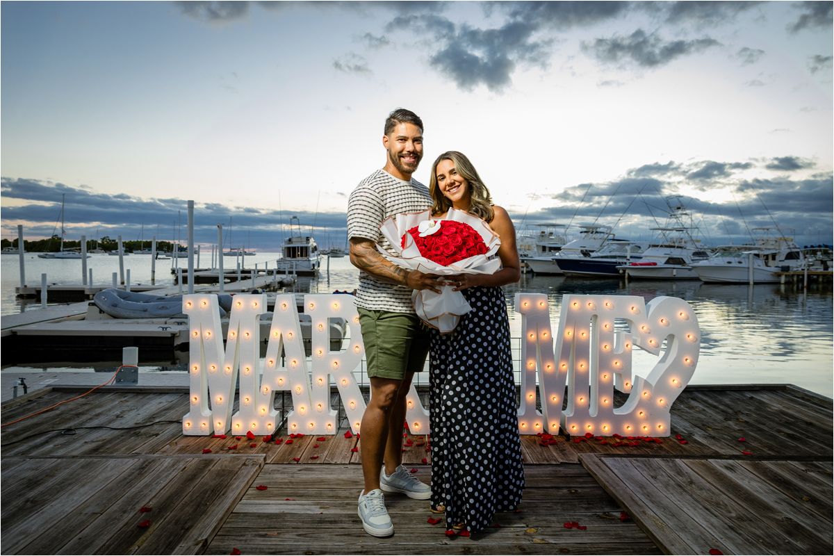 A couple stands on a dock at sunset with lighted WAR letters displayed behind them on the waterfront.