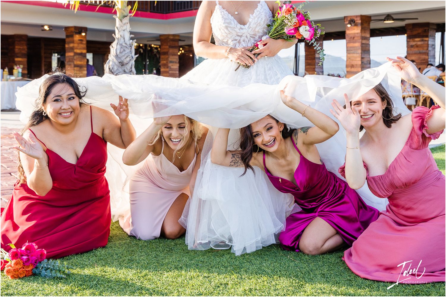Wedding party in pink and burgundy dresses poses playfully under bride's dress on lawn.
