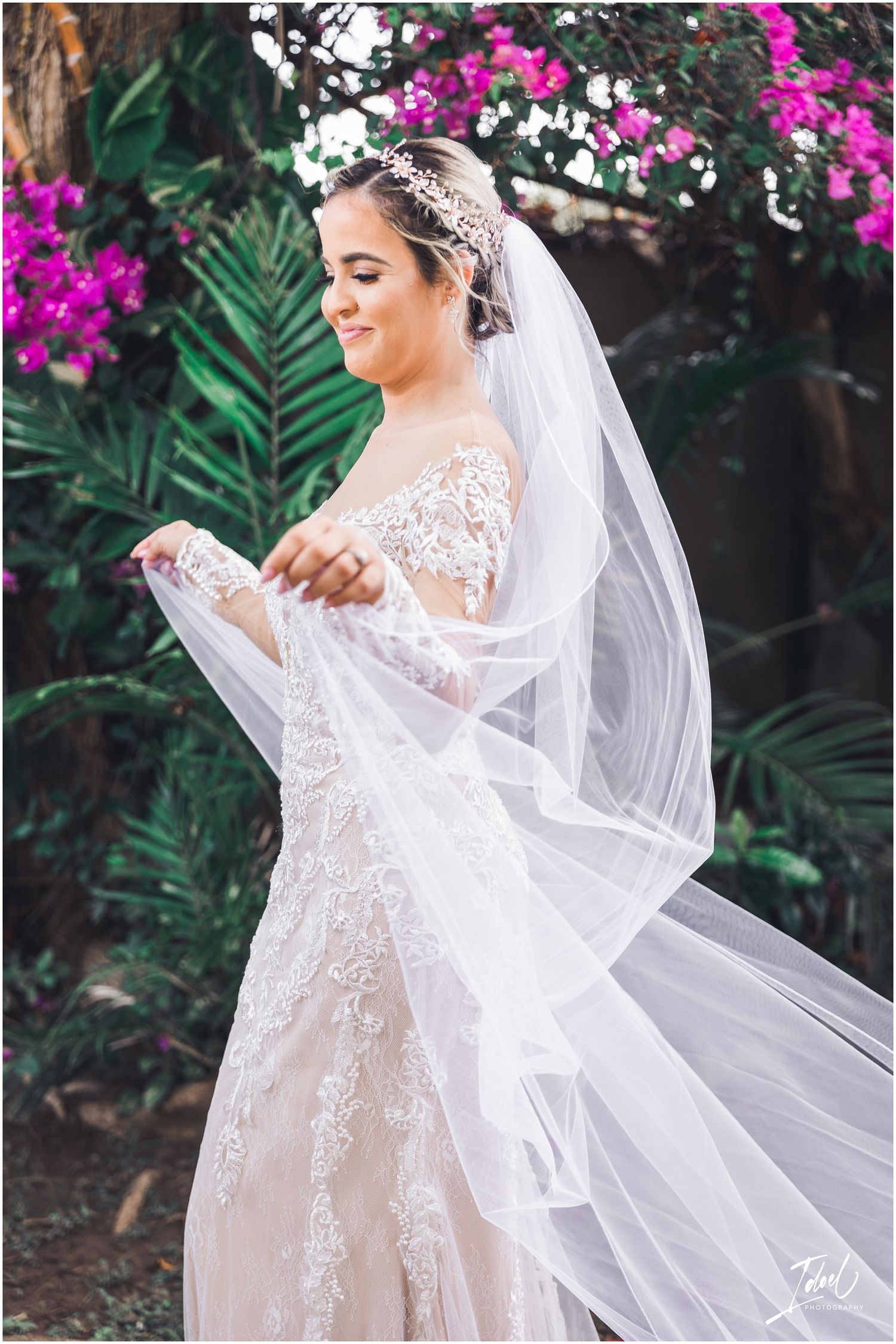 A bride in a delicate lace gown playfully holds her veil amidst blooming flowers.
