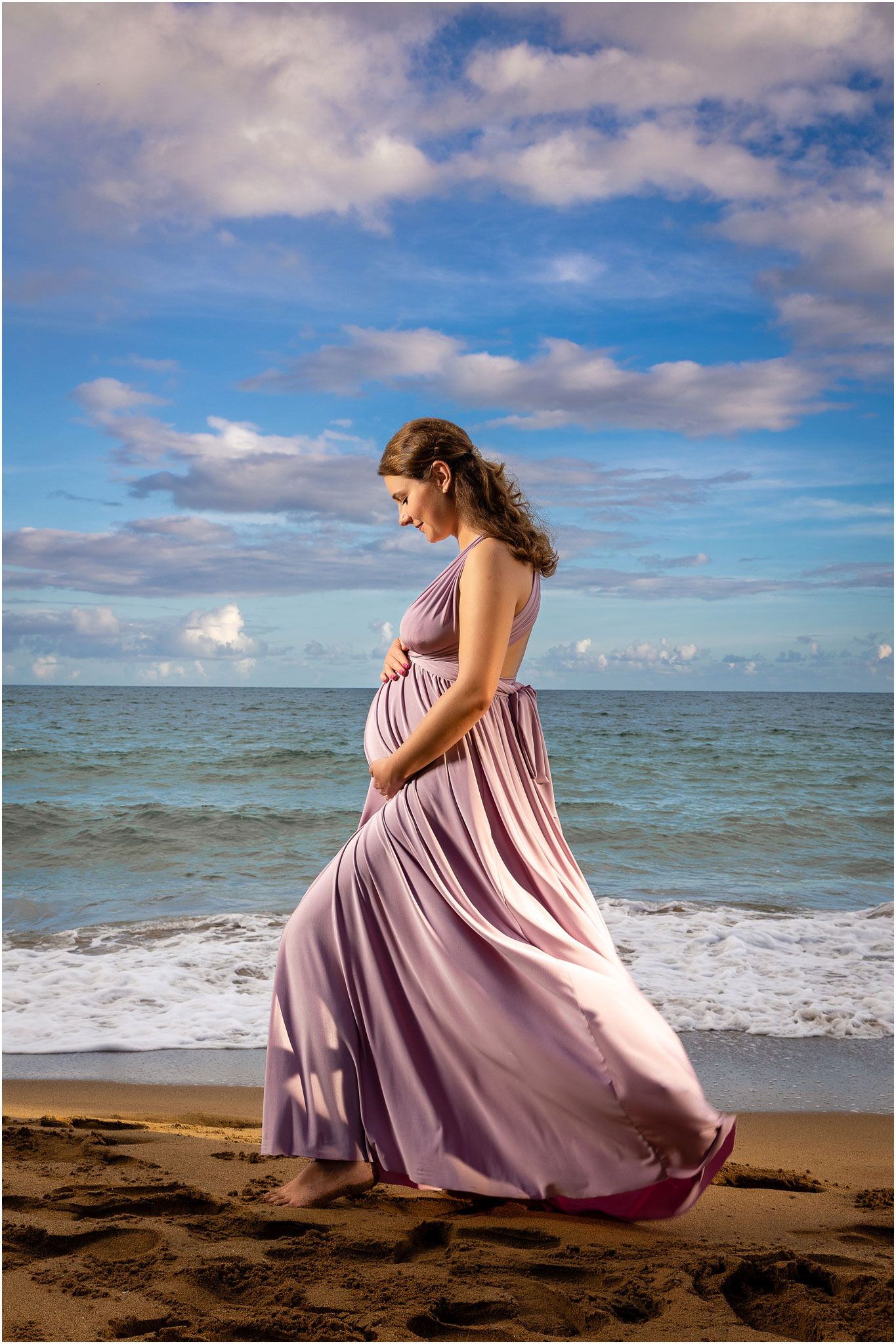 Flowing mauve dress billows in ocean breeze during beach maternity photoshoot.