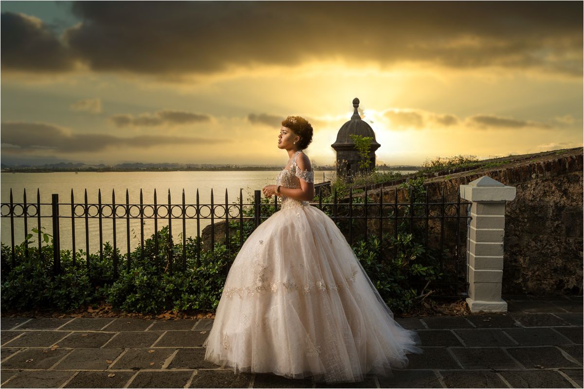A bride in a flowing white gown stands against a dramatic sunset sky overlooking a coastal landscape.