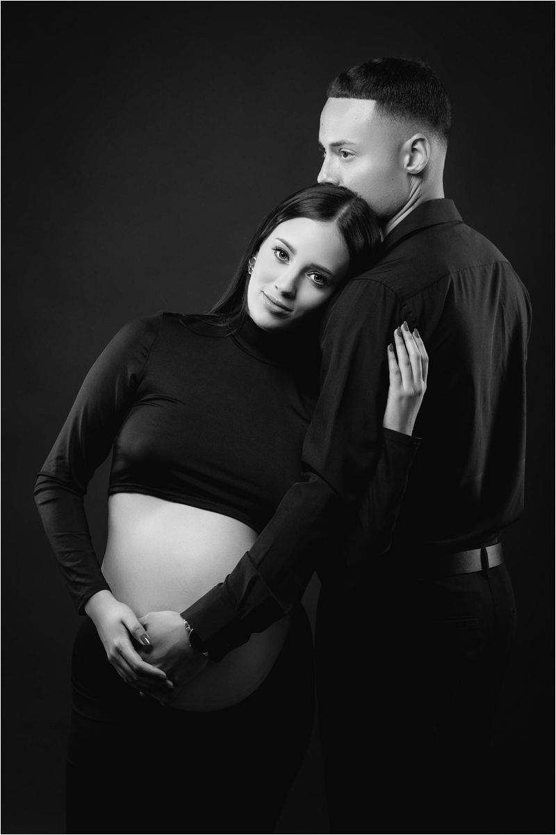 Artistic black and white maternity portrait showing an intimate moment between expectant parents in dark clothing.