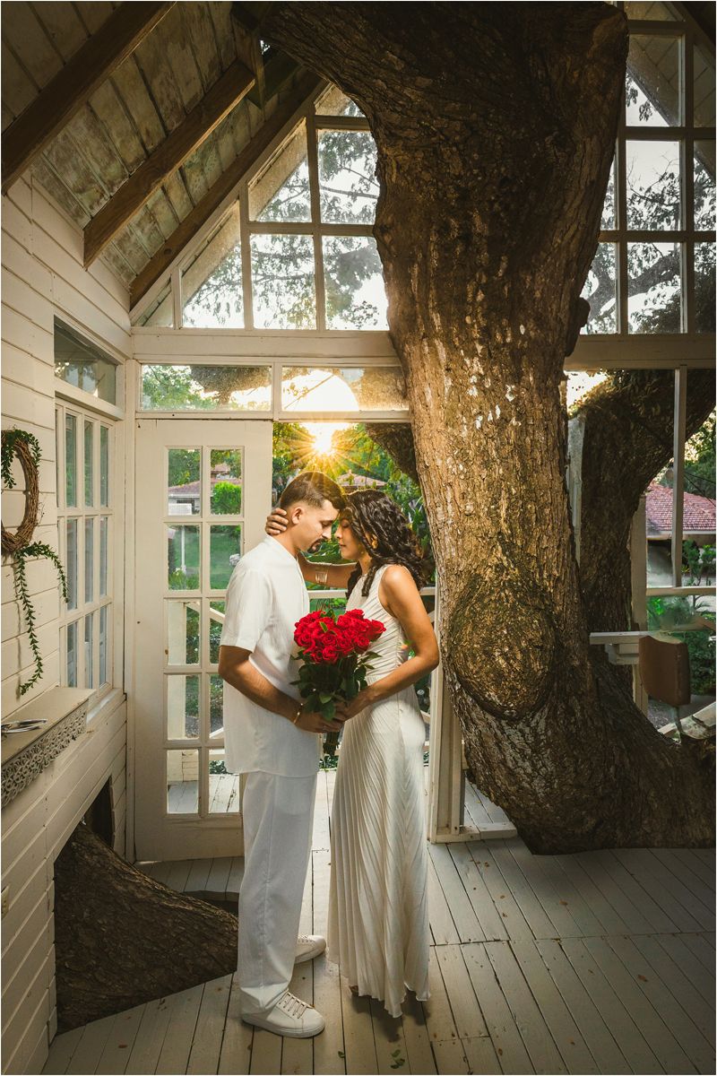 A romantic wedding couple poses together on a rustic wooden porch with a large tree trunk and sunlight streaming through windows.