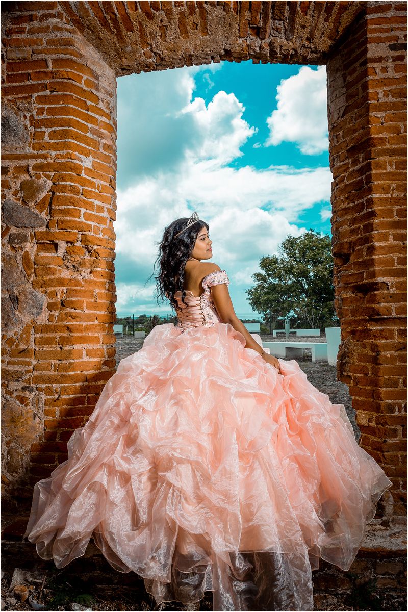 A stunning quinceañera dress in peach ruffles photographed against rustic brick columns under a bright blue sky.