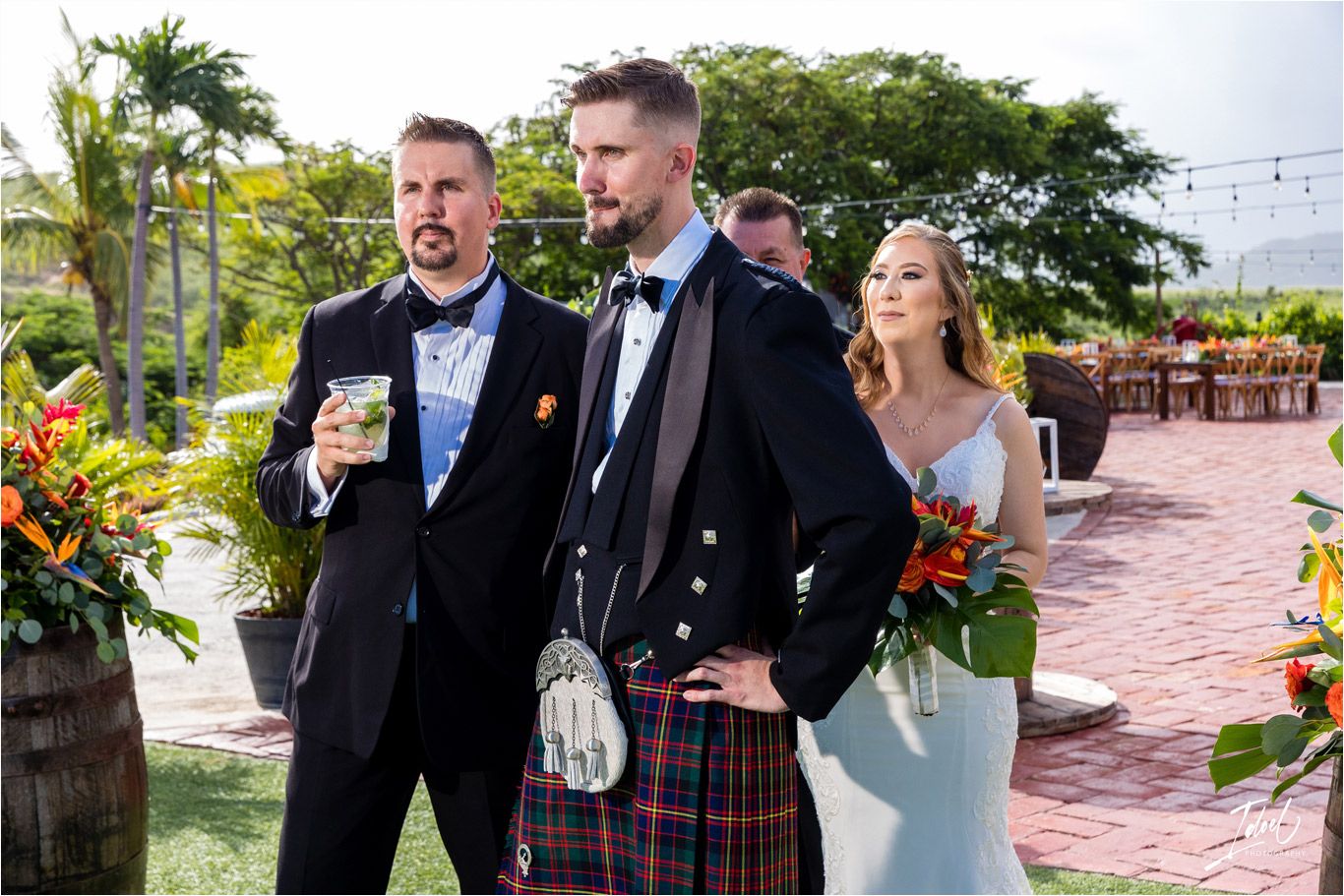 Wedding party in kilts and formal attire posing on an outdoor patio with tropical plants.