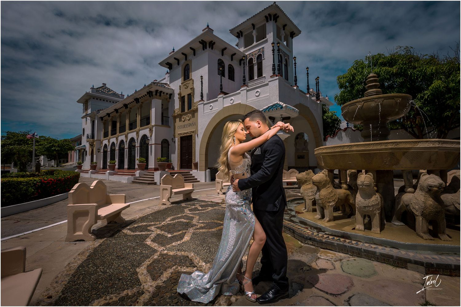 Couple in formal wear embracing outside historic building with dramatic cloudy sky.
