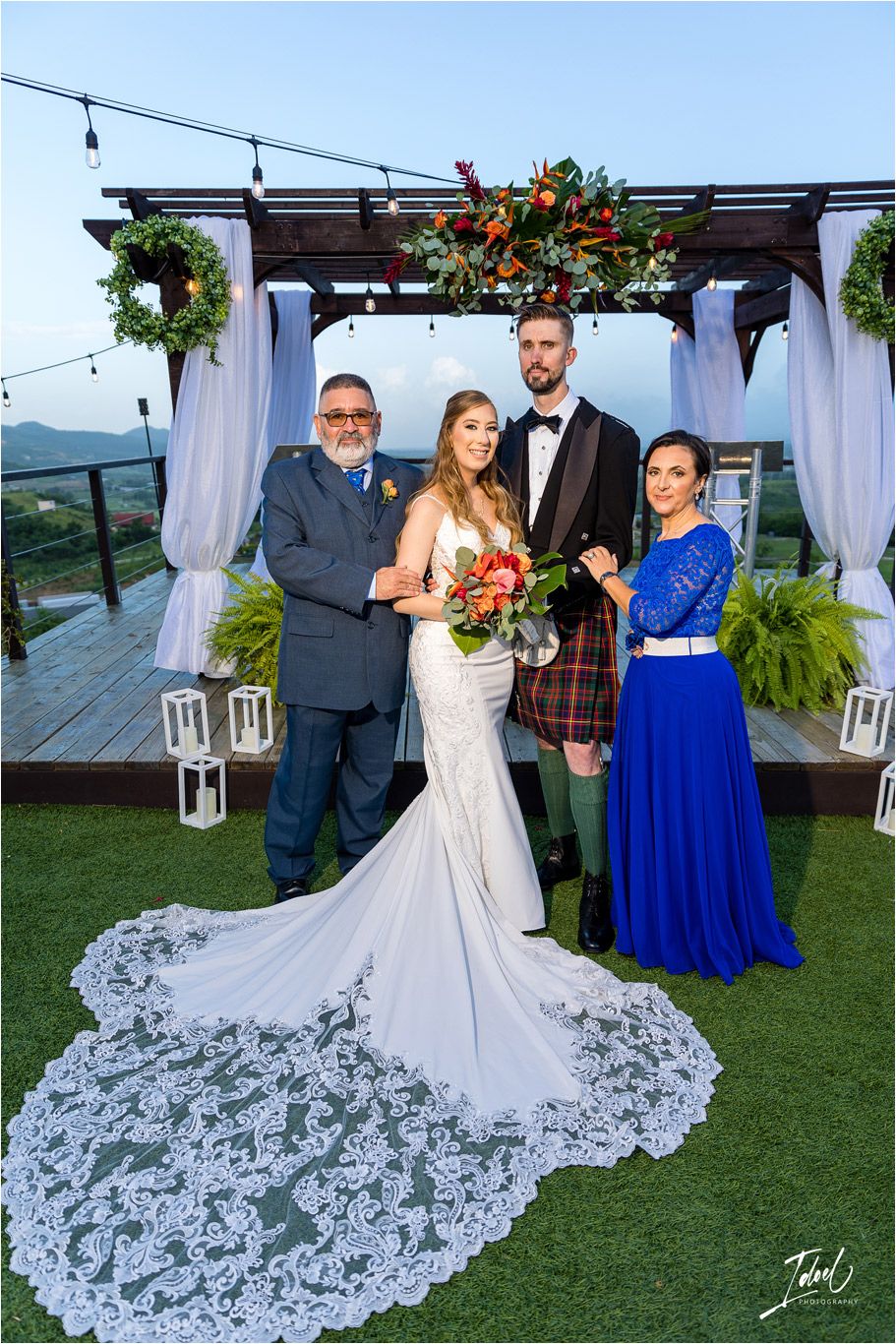 Bride in lace train gown poses with wedding party under decorated pergola with scenic mountain backdrop.