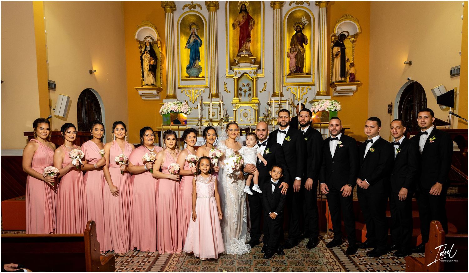 Full wedding party in pink dresses and black tuxedos poses together in front of church altar.