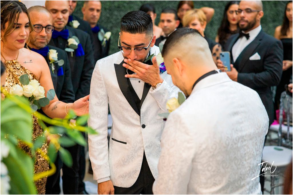 Two people in white tuxedo jackets share an emotional moment during an outdoor wedding ceremony.