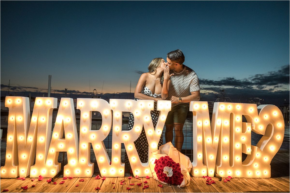 Illuminated MARRY ME marquee sign on wooden deck at sunset with bouquet of red roses.