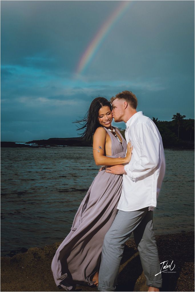 Romantic beach moment with couple embracing under a rainbow in the background.