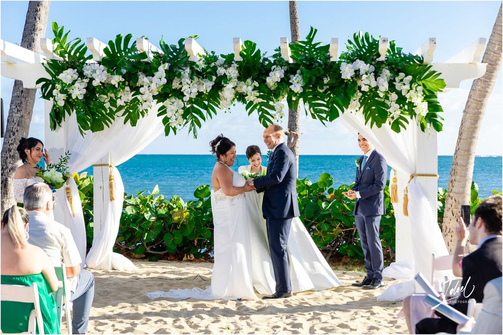 Wedding ceremony on beach with monstera leaf arch, white draping, and turquoise ocean views.
